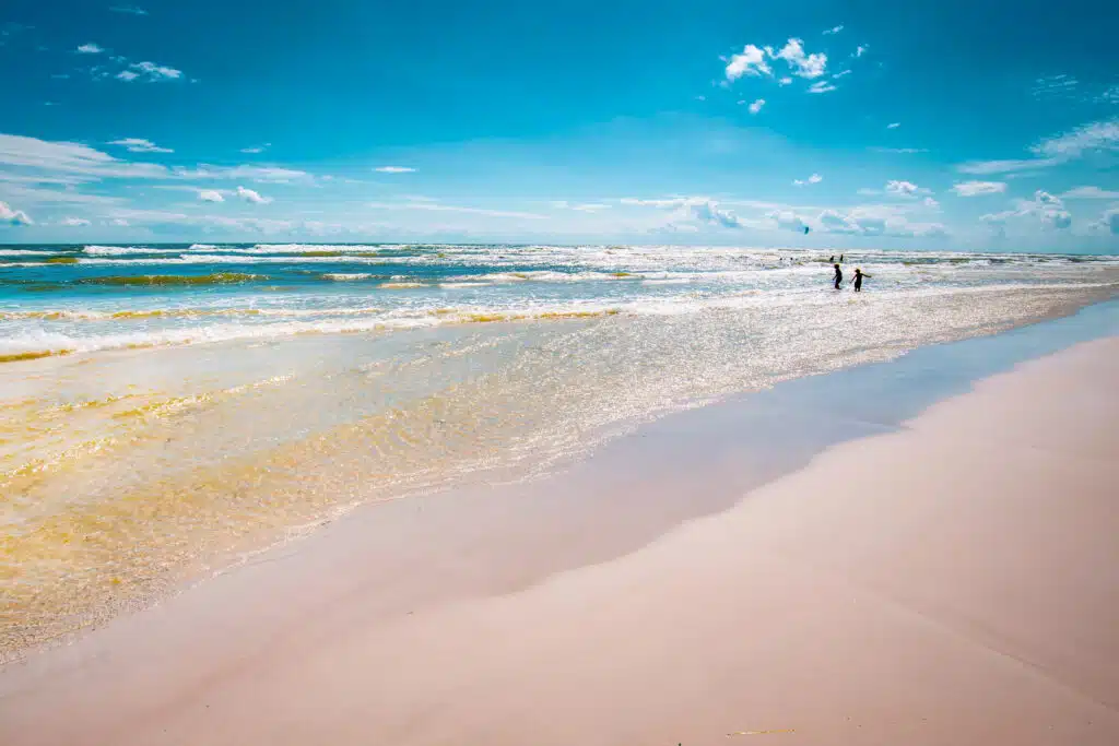 Szeroka piaszczysta plaża z płytką, przejrzystą wodą, w oddali dwie osoby w falach i latawce kitesurferów