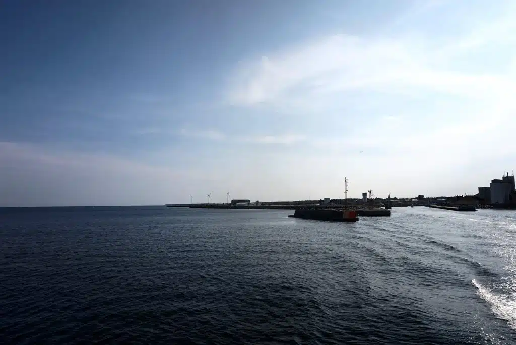 Wejście do portu w Nexø widziane z morza, falochron, silosy i wiatraki, z prawej spieniony kilwater