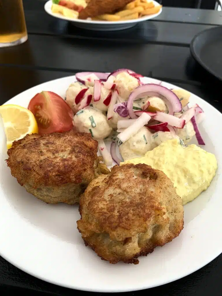 Dwa kotlety rybne fiskefrikadeller z remuladą, sałatką ziemniaczaną, pomidorem i cytryną na talerzu