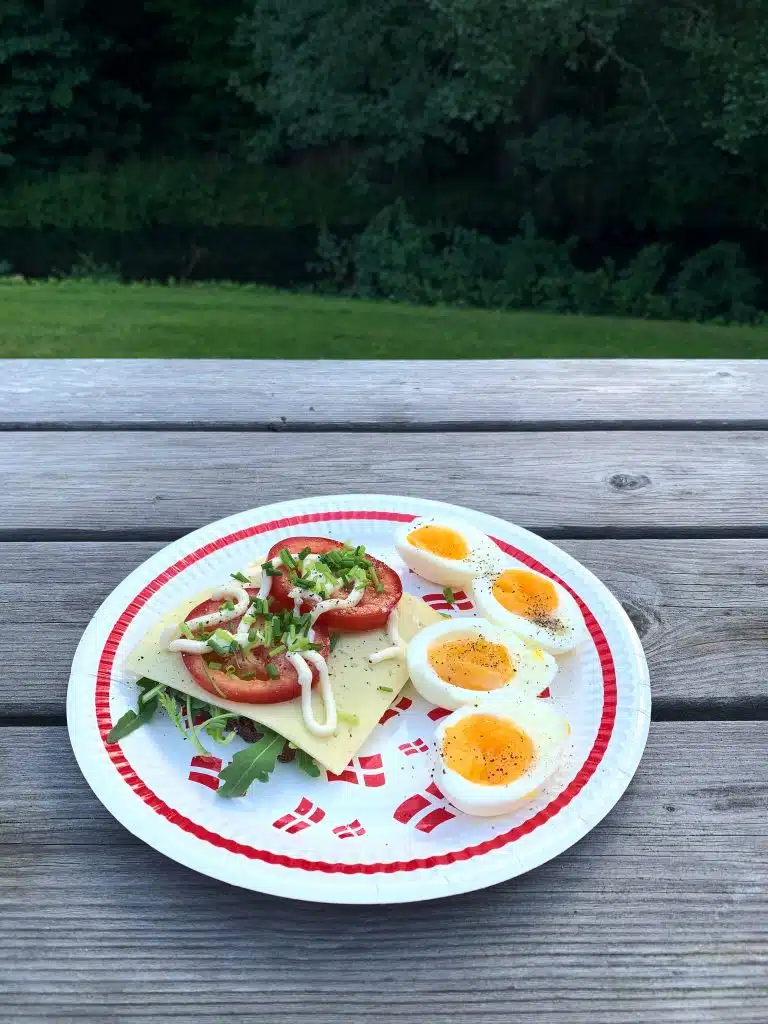 Kanapka z serem, pomidorem i rukolą oraz jajka na papierowym talerzu w duńskie flagi, na drewnianym stole z widokiem na trawnik i rzeczkę